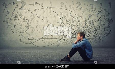 Upset and tired boy teenager sitting on the floor keeps hand to cheek looking thoughtfully and hopeless. Stressed student guy feels emotional discomfo Stock Photo