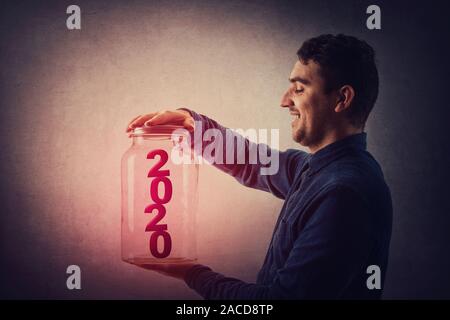Joyful young man opening the lid of a glass jar with red 2020 number inside. Start celebrating the new year. Happy holiday concept. Stock Photo
