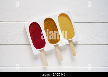 three different fruit smoothie popsicles in reusable silicone ice pop mold or form on white wooden background - making strawberry, passion fruit and banana flavor ice lollies Stock Photo