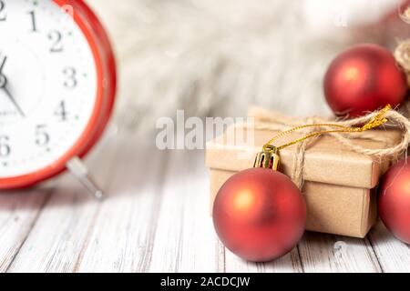 Gift box and blurred alarm clock on background on a wooden table decorated with a garland and red Christmas balls for the New Year or XMAS. Copy space Stock Photo