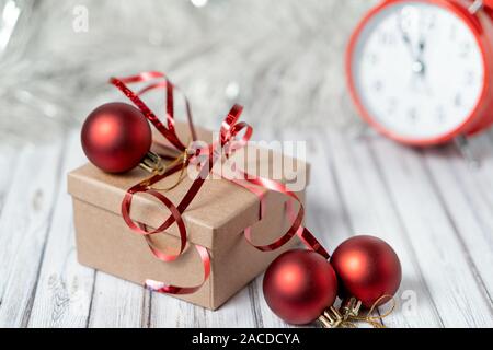 Gift box and blurred alarm clock on background on a wooden table decorated with a garland and red Christmas balls for the New Year or XMAS. Copy space Stock Photo