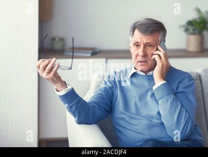 Senior Man Having Unpleasant Phone Conversation Sitting On Couch Indoor Stock Photo