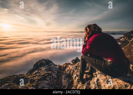 woman sitting on mountain peak watching sunrise over a sea of fog Stock Photo