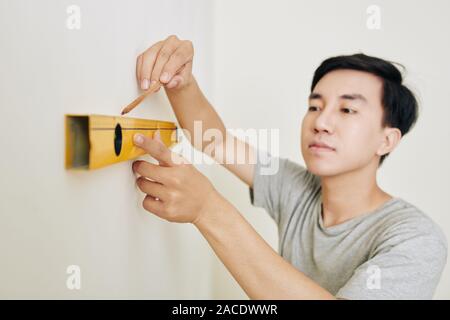 Handsome young Vietnamese man using spirit level when drawing stright line on the wall Stock Photo