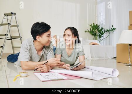 Beautiful young Vietnamese couple with tablet computer lying on the floor and discussing apartment plan Stock Photo