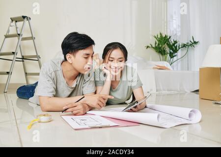 Happy young Vietnamese couple with blueprint and tablet computer lying on floor in their new house and discussing design ideas Stock Photo