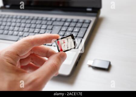 SD card in hand close-up. Laptop computer and card reader on desk in background Stock Photo