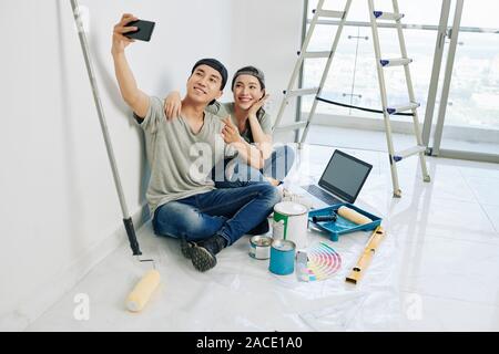 Happy young Vietnamese couple posing for selfie with tools and paints before renovating house Stock Photo
