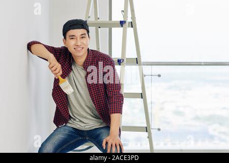 Portrait of young smiling Vietnamese man in plaid shirt with brush in hand leaning on wall and looking at camera Stock Photo