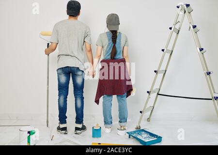 Young couple holding hands and looking at wall they are going to paint, view from the back Stock Photo