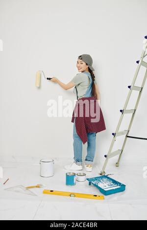 Happy young Vietnamese woman painting room walls with roller and smiling at camera Stock Photo
