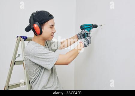 Young Asian man in protective ear muffs drilling hole in wall for hanging pictures Stock Photo