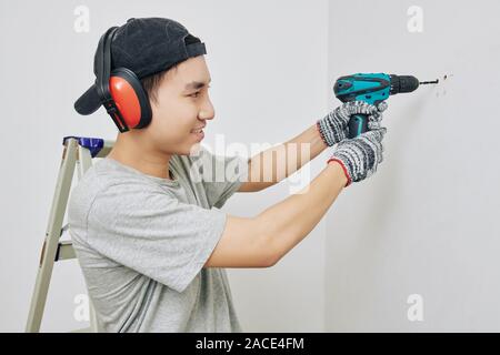 Smiling young Vietnamese man wearing protective ear muffs and gloves when drilling wall in apartment Stock Photo