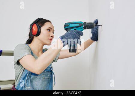 Pretty young Vietnamese woman concentrated on drilling wall in her room Stock Photo
