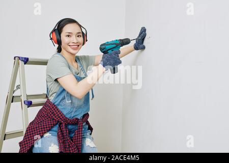 Portrait of pretty smiling young Asian woman wearing protective ear muffs when using electric drill Stock Photo