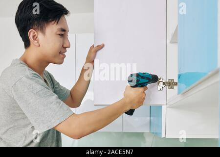 Handsome young Vietnamese man using electric screwdriver when assembling cupboard in kitchen Stock Photo