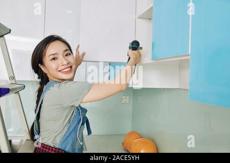 Cheerful beautiful Vietnamese young woman using electric screwdriver when fixing cupboard door in her kitchen