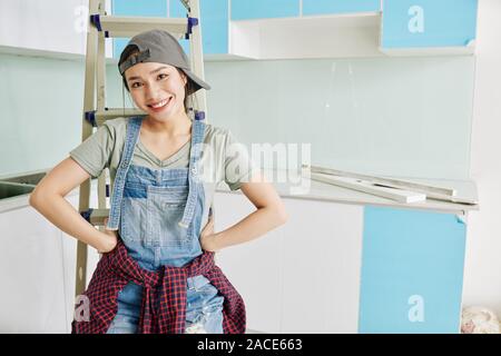 Portrait of happy young Asian woman in cap and denim overall standing in kithen she is renovating Stock Photo