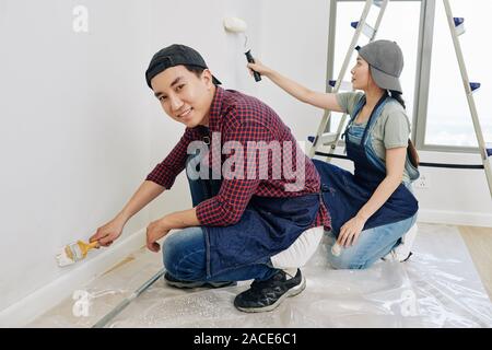Cheerful young Asian man painting wall with small brush when his girlfriend using foam roller Stock Photo