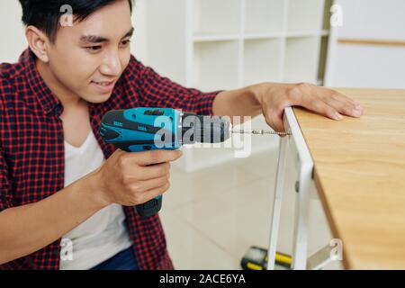 Positive excited handsome young Asian man using screwdriver for fixing table surface in place Stock Photo