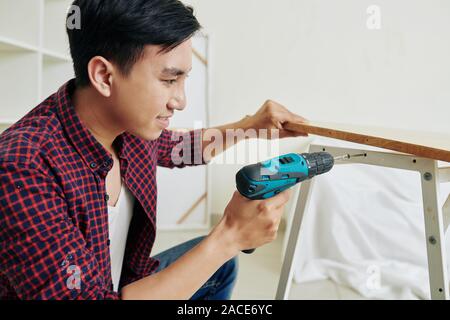 Smiling young Vietnamese man using electric screwdriver when accembling bench at home Stock Photo