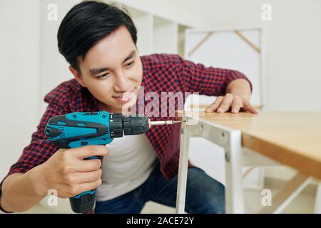 Positive young Vietnamese man assembling furniture in new apartment and tightening screw with electric screwdriver Stock Photo