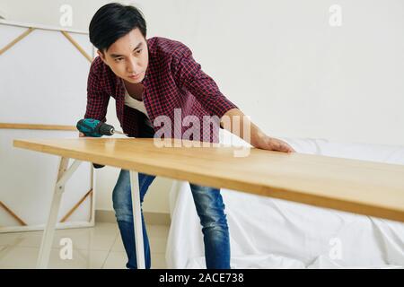 Serious Vietnamese young man with electric screwdriver assembling table in new apartment Stock Photo