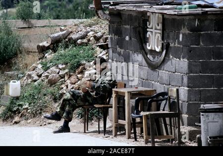 11th August 1993 During the Siege of Sarajevo: a Bosnian-Serb soldier relaxes outside his bunker on Mount Trebevic, above Sarajevo. A belt of spent M2 Browning .50 calibre cartridges hang below a Serbian cross symbol on the front of the hut. Stock Photo