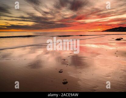 Charmouth, Dorset, UK. 2nd December 2019. UK Weather: Spectacular colours are reflected in the wet sand at low tide as the sun sets over the beach at the coastal resort of Charmouth.  Footprints in the wet sand were the only sign of life on the deserted beach.  Credit: Celia McMahon/Alamy Live News. Stock Photo