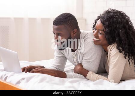 Cheerful afro spouses lying in bed using laptop together Stock Photo