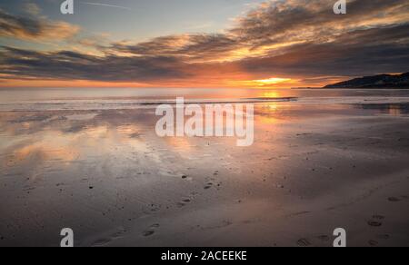 Charmouth, Dorset, UK. 2nd December 2019. UK Weather: Spectacular colours are reflected in the wet sand at low tide as the sun sets over the beach at the coastal resort of Charmouth.  The popular beach is deserted as the sun drops below the horizon towards Lyme Regis on what has been a bright and cold winters day.   Credit: Celia McMahon/Alamy Live News. Stock Photo