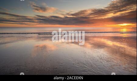 Charmouth, Dorset, UK. 2nd December 2019. UK Weather: Spectacular colours are reflected in the wet sand at low tide as the sun sets over the beach at the coastal resort of Charmouth.  The popular beach is deserted as the sun drops below the horizon towards Lyme Regis on what has been a bright and cold winters day.   Credit: Celia McMahon/Alamy Live News. Stock Photo