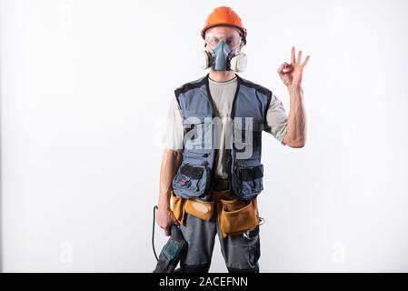 Builder with a drill puncher, in a helmet, and a respirator. Shows okay sign Stock Photo