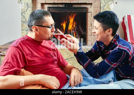 Married mature couple are sitting on bean bag chairs and drinking wine near fireplace while celebrating event. Husband and wife celebrates date at the Stock Photo