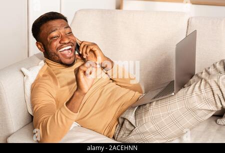 Cheerful Guy Talking On Phone Lying With Laptop On Couch Stock Photo