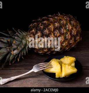 Sliced pineapple on old rustic desk with knife and with whole pineapple behind. Black background. Stock Photo
