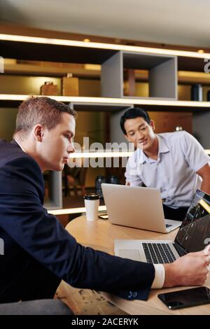 Serious businessman checking stock market data on laptop screen when meeting with Vietnamese colleague in cafe Stock Photo