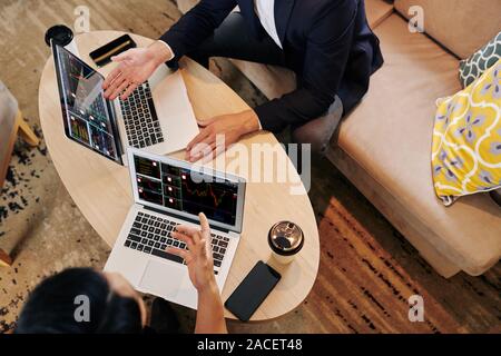 Financial analysts sitting at cafe table with opened laptops and discussing charts with sales data on screens, view from above Stock Photo