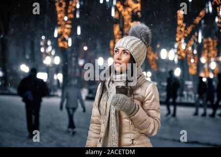 Handsome girl in gray knitted hat with pom pom, scarf and gloves, pink jacket standing on street in evening. Beautiful lady holding coffee cup and loo Stock Photo