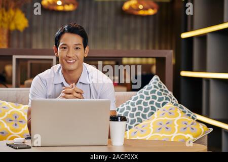 Portrait of smiling handsome Asian freelancer sitting at table in cafe, working on laptop and looking at camera Stock Photo