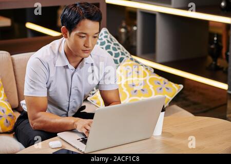 Young serious Vietnamese businessman spending work day in cafe, he is looking at computer screen when typing on keyboard Stock Photo
