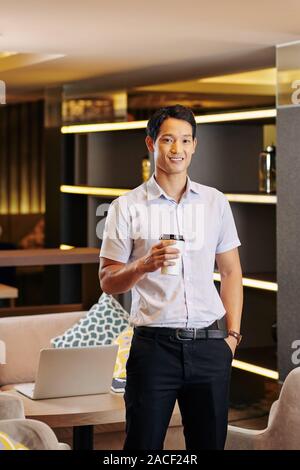 Young positive Vietnamese entrepreneur drinking cup of tasty coffee from disposable cup Stock Photo