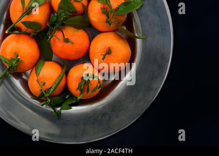Fresh orange mandarins with green leaves lie on an old metal plate in front of a dark background Stock Photo