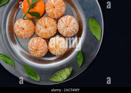 Fresh peeled tangerines with green leaves lie on an old metal plate against a dark background Stock Photo