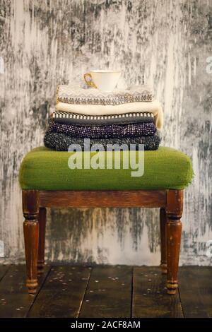 Pile of knitted warm grey and white blankets, scarves and woolen sweaters for winter or fall cold weather on white stool near white and gray wall and Stock Photo