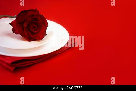 Romantic table setting for Valentines day or dinner date celebration wedding. Valentines day table setting with red rose bud, white plates on red tablecloth background. space for text Stock Photo