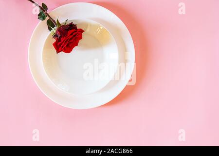 Romantic table setting for Valentines day or dinner date celebration wedding. Valentines day concept with red rose bud white plates on pastel pink tablecloth background. Flat lay, top view copy space. Stock Photo