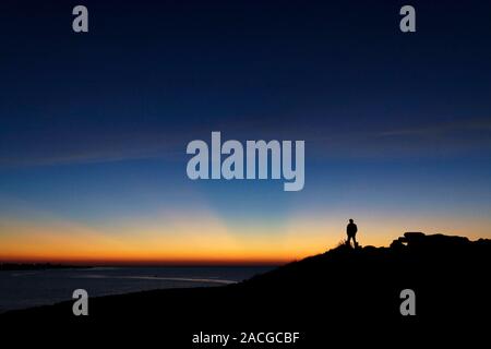 Sun beams. Person on a rock watching orange crepuscular rays emerging from behind the western horizon after sunset. These rays of sunlight are streami Stock Photo