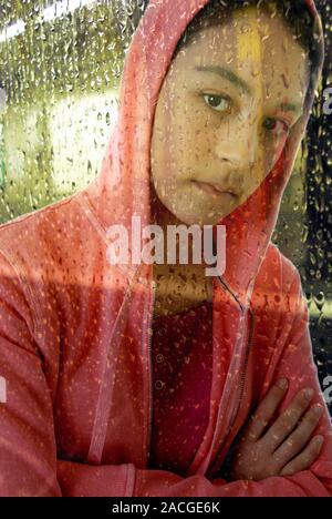 Woman in a hoodie. Young woman wearing a hooded jumper, behind a window pane covered with raindrops. Stock Photo