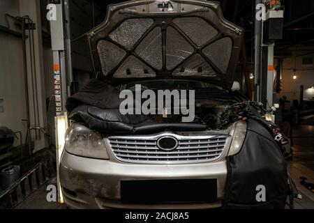 Disassembled gray car in the workshop raised on the lift prepared for repair with opened hood, engine removed, internal equipment, parts  visible. Aut Stock Photo